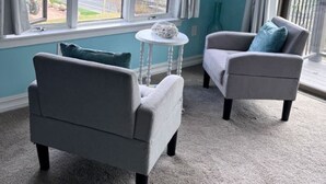 A room with two grey armchairs, a small round table, and a view of a road and trees through large windows. - Spacious House for Beach Getaway - Outdoor Shower -Beach Pass Included! (Charlestown)