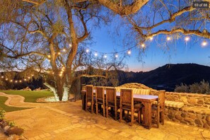 An outdoor dining area with a stone wall, wooden chairs, and a table under string lights.