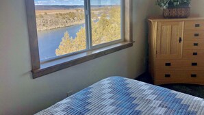 Un dormitorio con un ventanal grande que ofrece vistas a un río y árboles, un aparador de madera y una cama con colcha estampada.