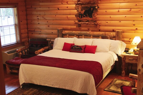 A log cabin room with a large bed, a chair, a wooden headboard, and a window with blinds.