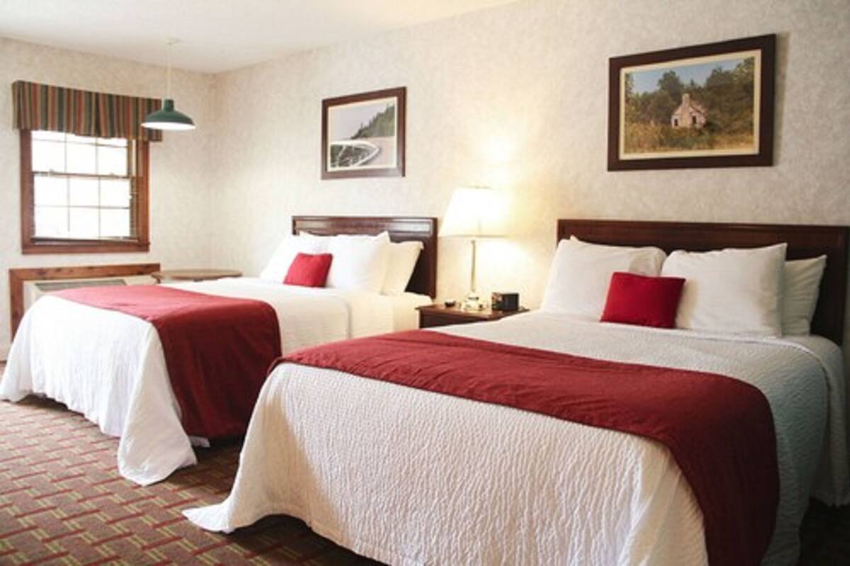 Quarto de hotel com duas camas, cada uma com colcha vermelha e branca, cabeceira de madeira, mesa de cabeceira com abajur e janela com vista para uma paisagem.