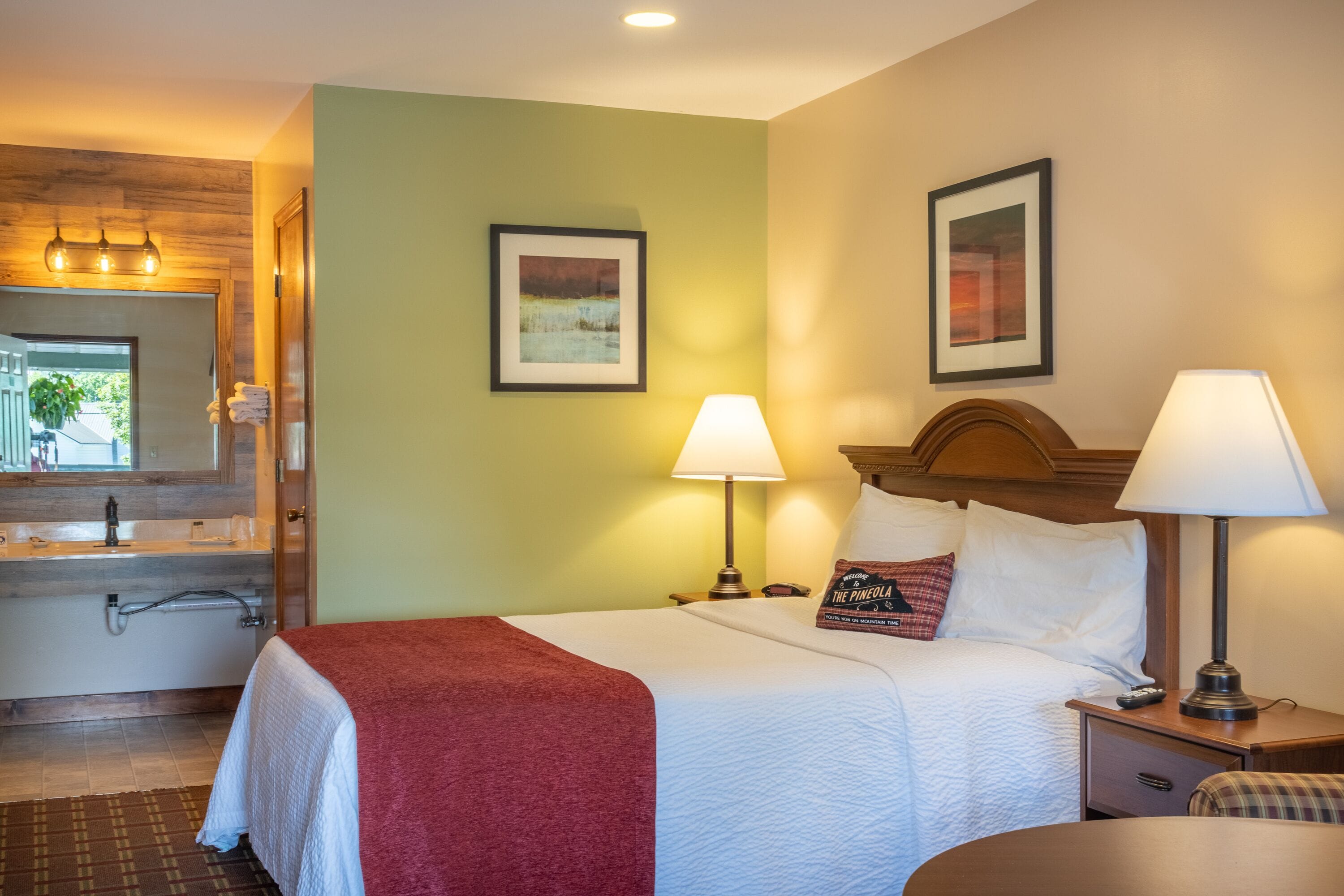 A hotel room with a bed, two bedside lamps, a nightstand, a bathroom with a sink and mirror, and two framed pictures on the wall.