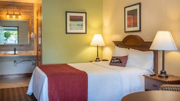 A hotel room with a bed, two bedside lamps, a nightstand, a bathroom with a sink and mirror, and two framed pictures on the wall.