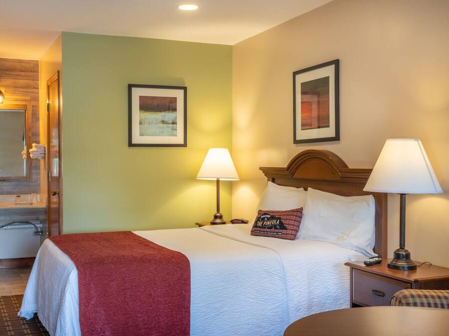 A hotel room with a bed, two bedside lamps, a nightstand, a bathroom with a sink and mirror, and two framed pictures on the wall.