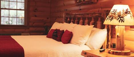 A wooden cabin bedroom with a bed, pillows, a nightstand, a lamp, and a window.