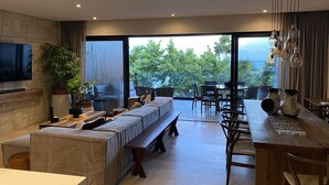 A modern living room with a large TV, comfortable seating, a dining area, and a view of greenery and the sea.