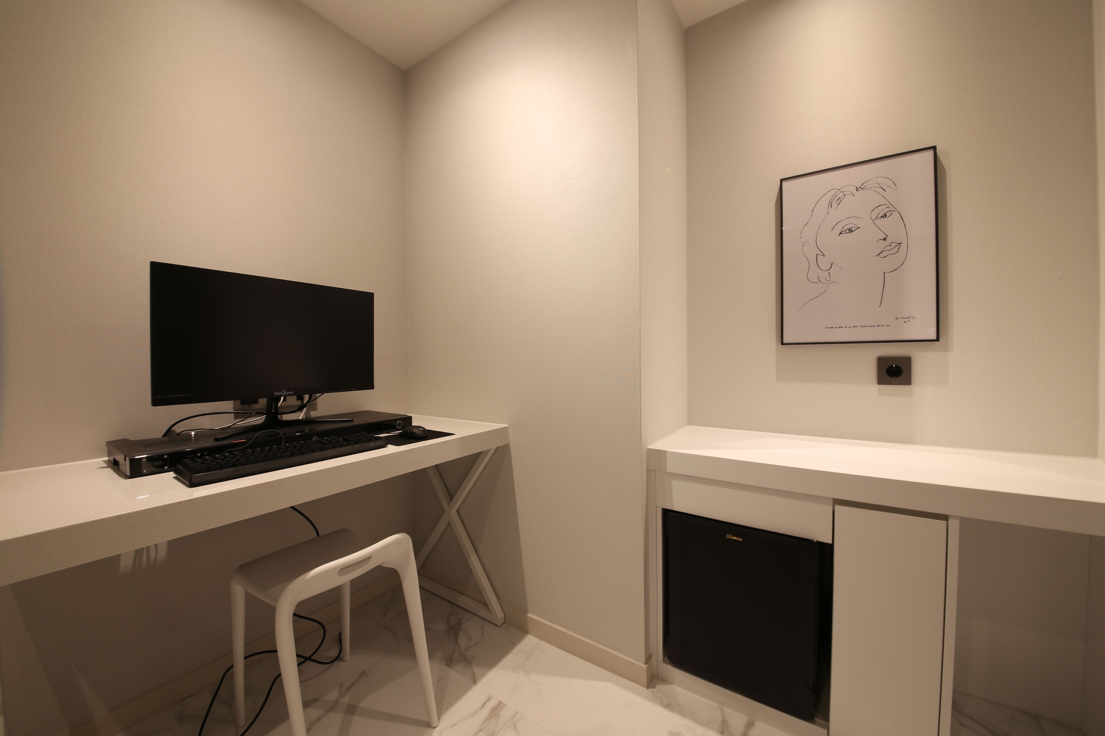 Un espacio de oficina moderno y minimalista con un escritorio blanco, monitor negro y teclado. Un dibujo enmarcado está en la pared, y hay una silla blanca frente al escritorio.