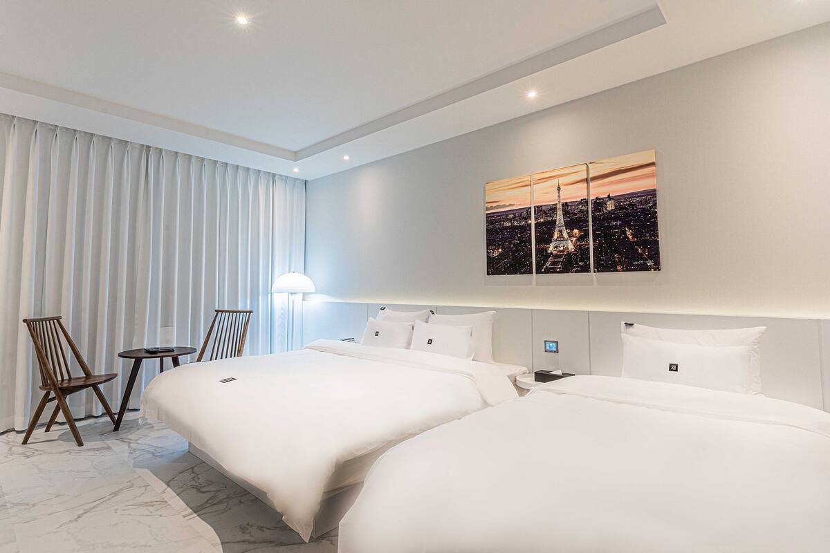 A hotel room with two beds, a desk, chairs, and a cityscape painting on the wall.