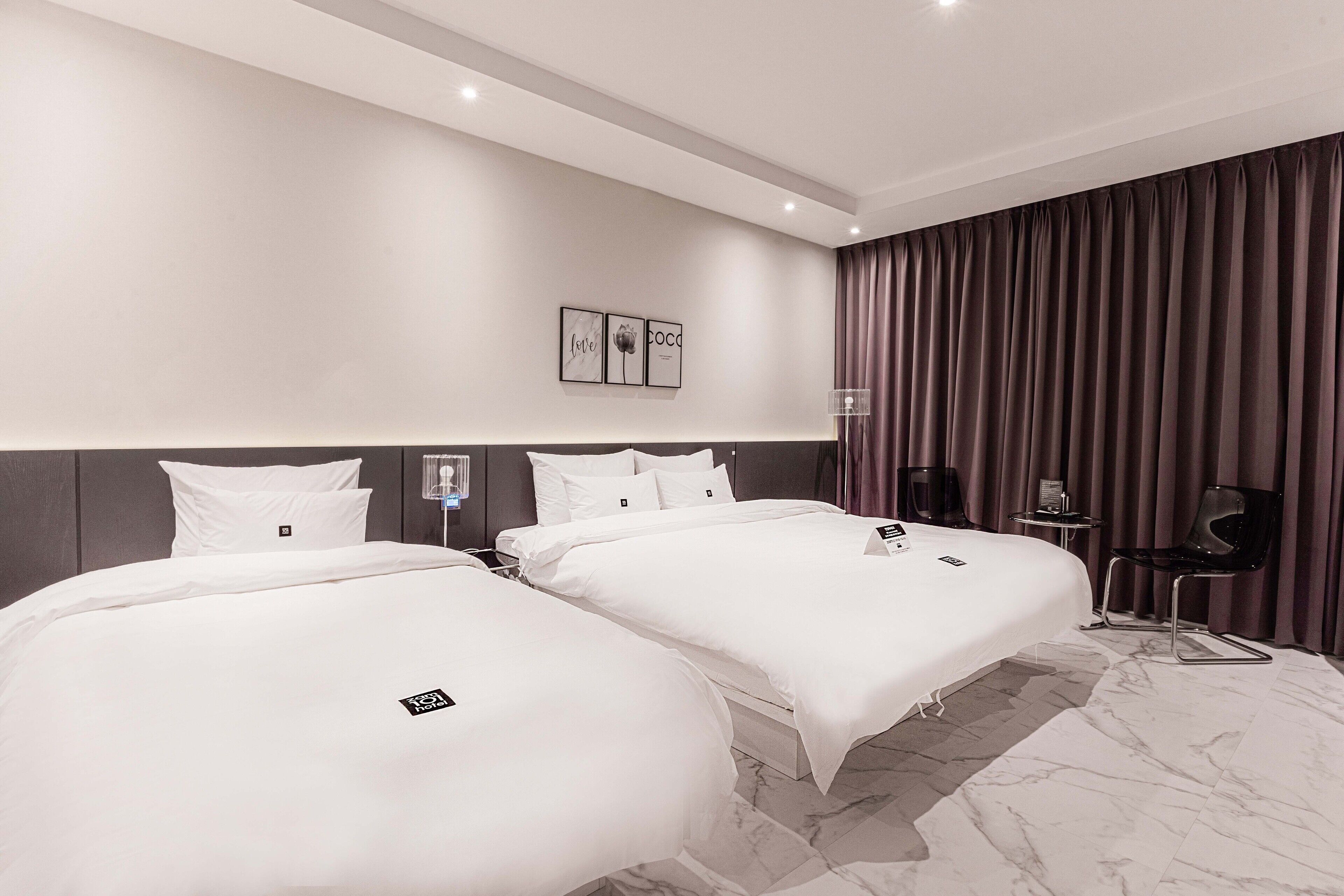 A hotel room with two beds, a marble floor, a large window with curtains, and a small table with a lamp.
