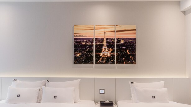 Ein Hotelzimmer mit einem großen Stadtbild, das den Eiffelturm zeigt, einem Bett mit weißer Bettwäsche und einem Nachttisch mit einer Lampe.