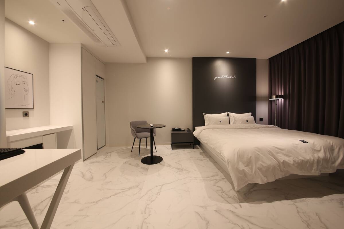 A modern hotel room with a marble floor, a bed with white bedding, a desk, a chair, a bedside table, and a wall-mounted artwork.