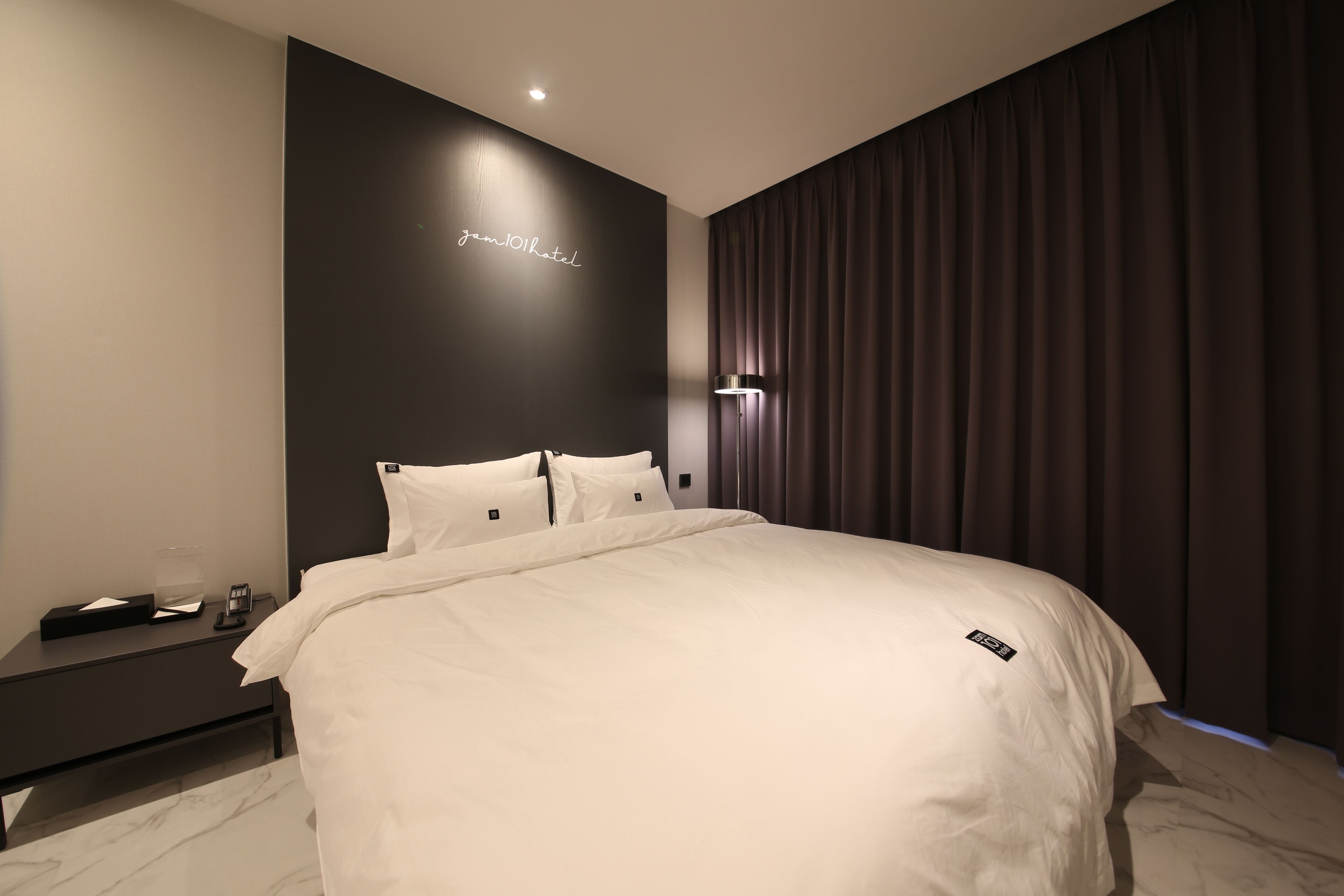 A modern hotel room with a large bed, a nightstand with a lamp, and a dark wall with a neon sign.