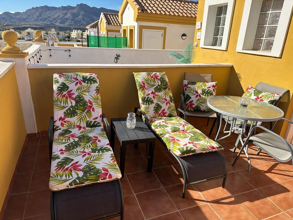 A terrace with two lounge chairs and a small table, surrounded by yellow walls and a view of mountains.