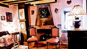 Un salon chaleureux avec une cheminée, deux fauteuils à carreaux rouges et blancs, un canapé et une table recouverte d’une nappe à motifs floraux.