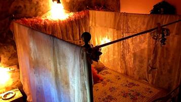 Een kamer met een bed dat bedekt is door een doorschijnende, bloemenpatroon-hemelbed. Een klein tafeltje met een lamp is links zichtbaar.