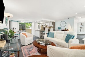 A modern living room with a white sofa, a glass coffee table, and a kitchen in the background.