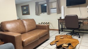 Une pièce avec un canapé en cuir marron, une table basse en bois, un bureau avec un ordinateur, une chaise et un téléviseur fixé au mur.