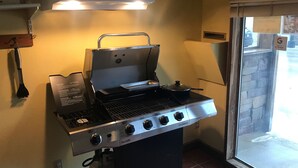Un barbecue à gaz doté de quatre brûleurs et d’un couvercle, situé dans une cuisine avec une fenêtre et une porte.
