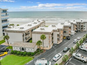 A multi-story hotel with balconies overlooking a beach, a parking lot with several cars, and a clear view of the ocean. - 🌊Wake Up to Ocean Views! Beachfront Balcony | Pool | Car Free Beach | Shore Villas (New Smyrna Beach)