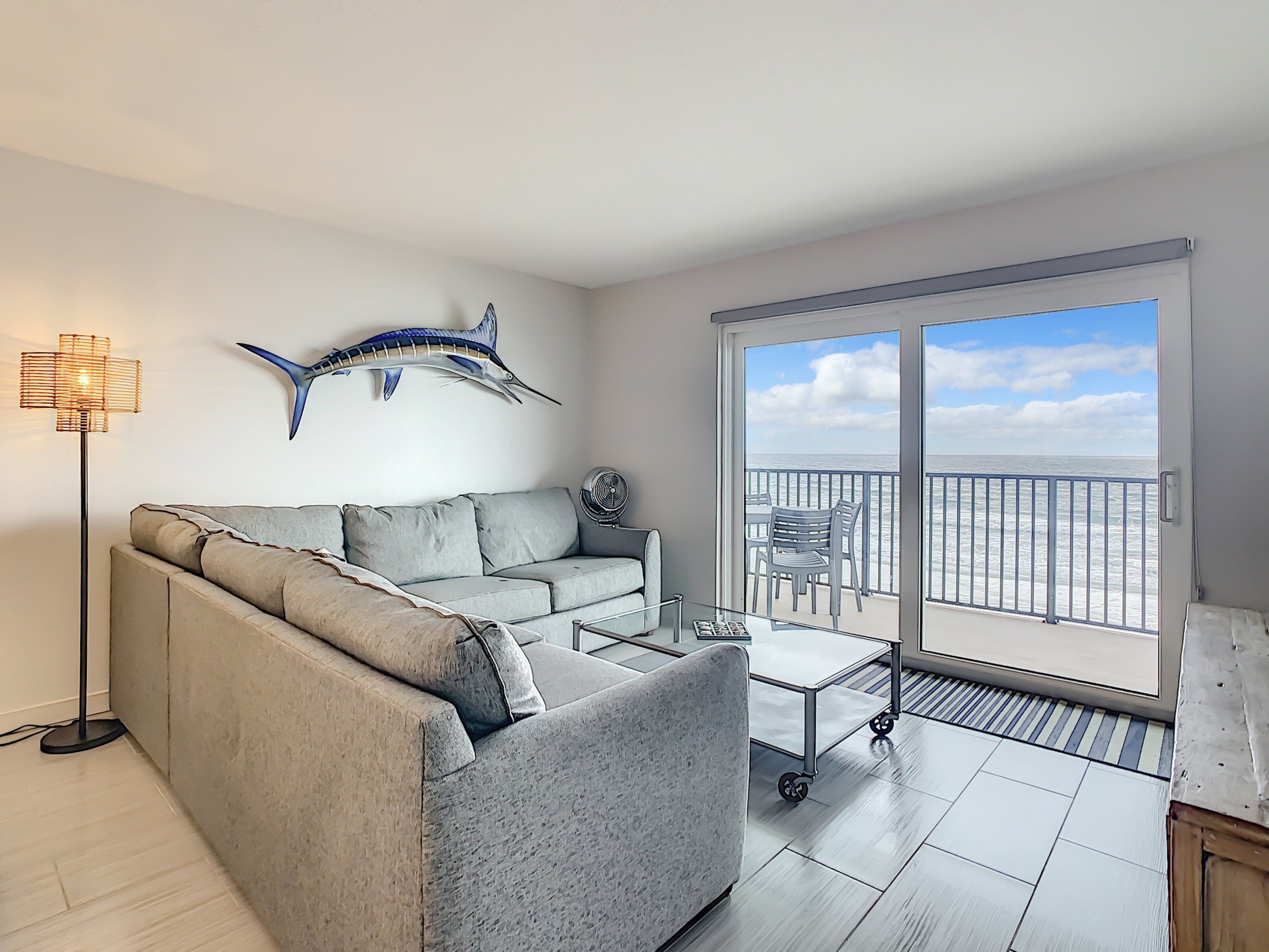 A modern living room with a grey sofa, a glass coffee table, and a large window overlooking the sea.