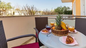 A table with breakfast items including fruits, pastries, and drinks on a balcony with wicker chairs and a red cushion.