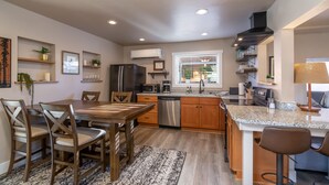 A modern kitchen with a dining area, wooden cabinets, stainless steel appliances, and a window with blinds. - Tumalo House (Bend)