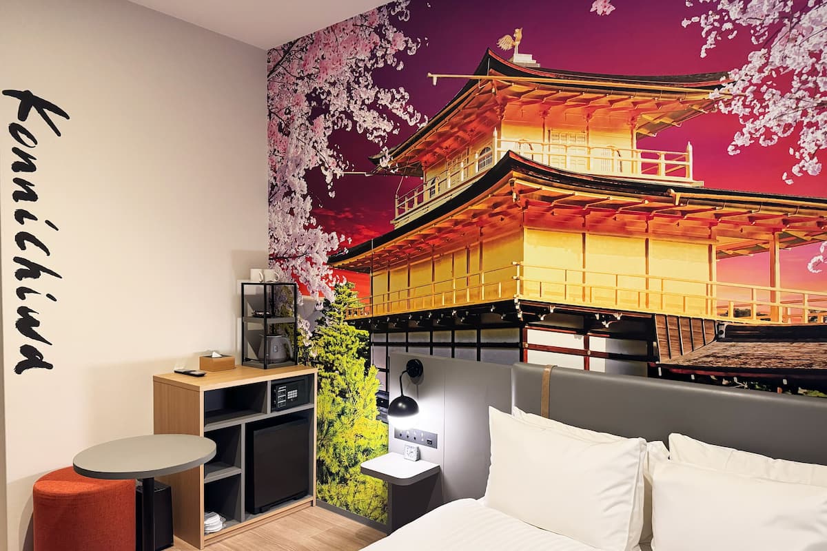 A hotel room with a bed, a desk, a chair, a small table, and a wall with a cherry blossom mural.