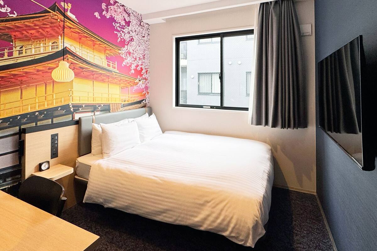A hotel room with a large bed, a small bedside table, a nightstand, a window with curtains, and a mural on the wall.