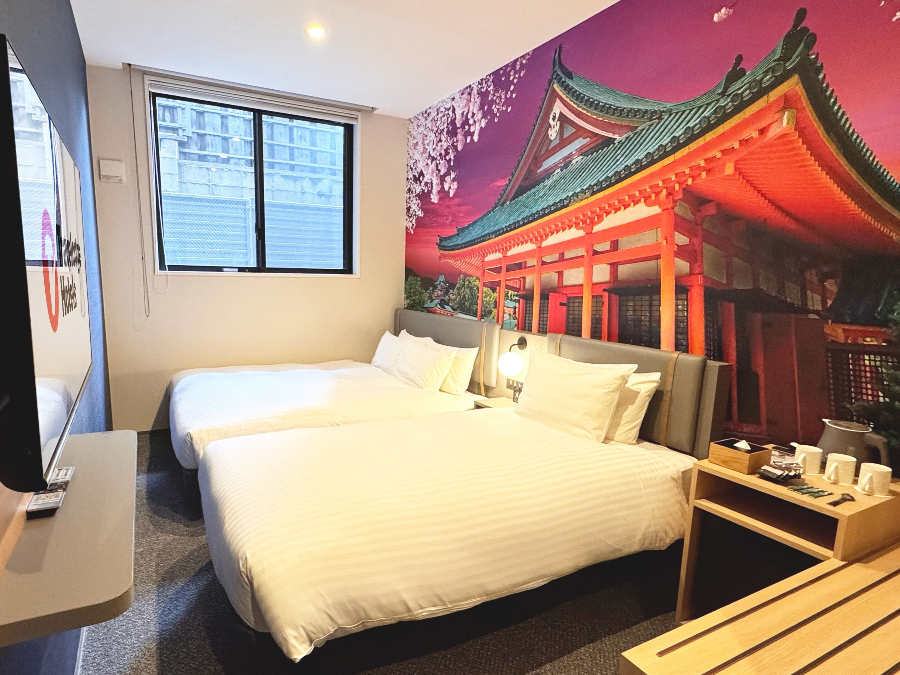 A hotel room with two beds, a TV, a window, and a mural of a traditional building.