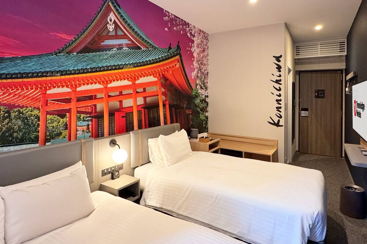 A hotel room with two beds, a nightstand with a lamp, a wall-mounted TV, and a mural of a traditional building.