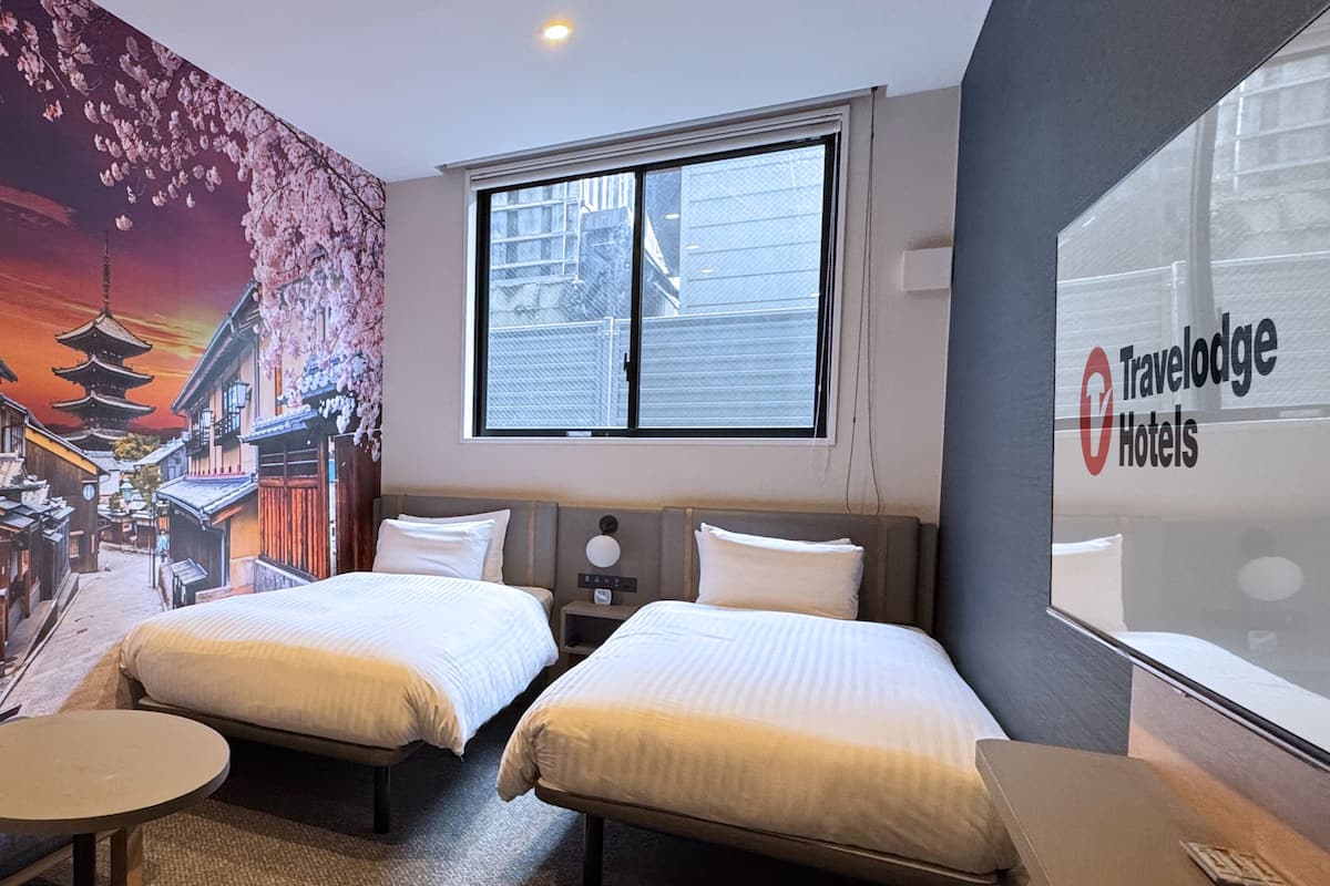 A hotel room with two beds, a small table, and a large window.