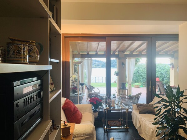 A living room with a sofa, a coffee table, and a TV stand. There is a sliding glass door leading to an outdoor patio area with a table and chairs. - The House by the Lake - Trevignano Romano (Trevignano Romano)
