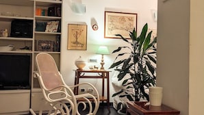 Un salon comprenant une bibliothèque, un téléviseur, un fauteuil à bascule, une table avec une lampe et une plante en pot.