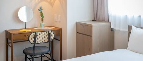 A hotel room with a wooden desk, a chair, a small potted plant, a bedside table, a bed, and a window with curtains.
