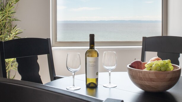Una mesa de comedor con una botella de vino, dos copas y un cuenco de fruta junto a una ventana con vistas al mar.