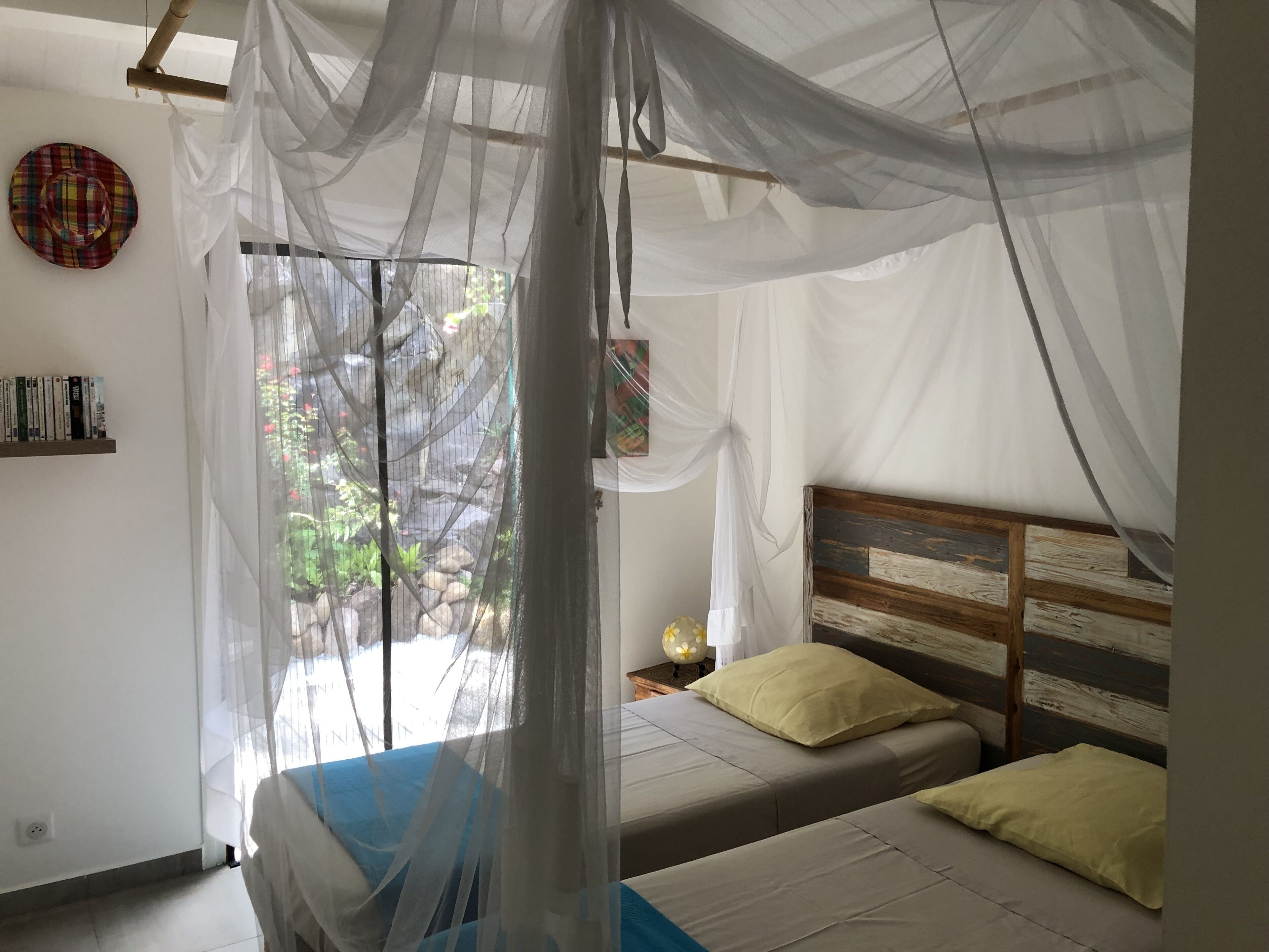 A bedroom with two beds, a mosquito net, a bedside table with a lamp, and a wall-mounted shelf with books.