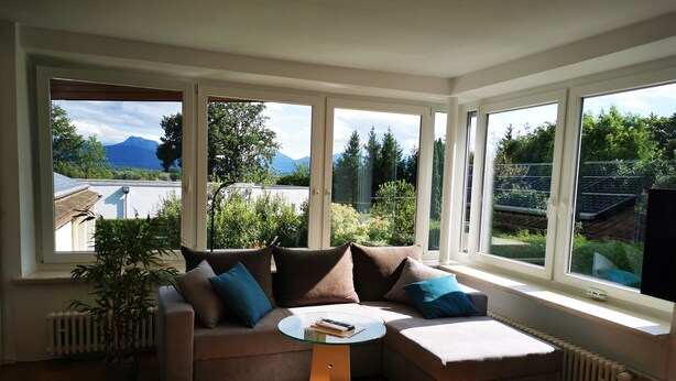 A cozy living room with a sectional sofa, a glass coffee table, and large windows offering a view of greenery and mountains.