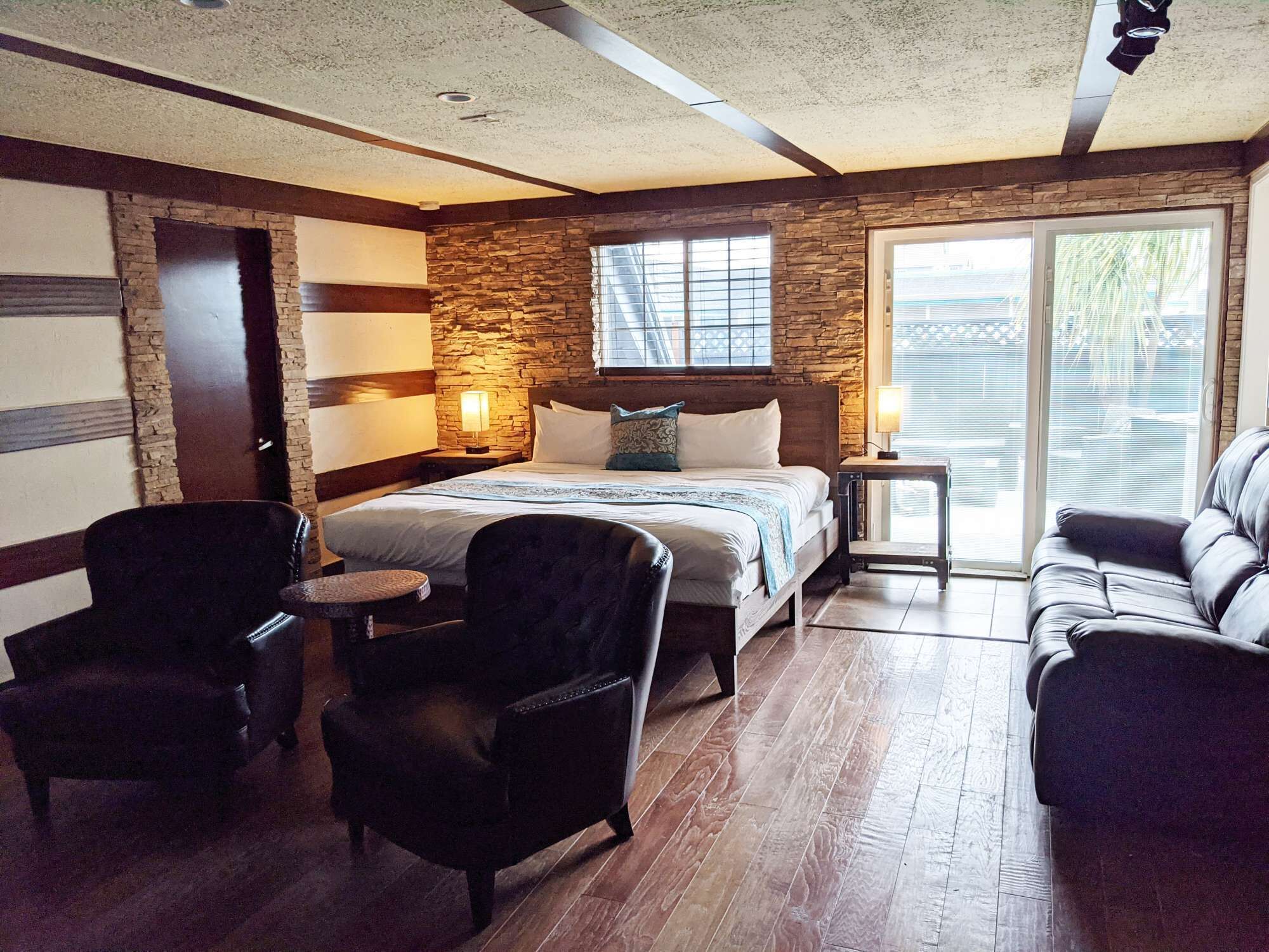 Une chambre d’hôtel comprenant un lit, deux fauteuils, une petite table, un mur décoratif en pierre et une porte-fenêtre coulissante donnant sur l’extérieur.