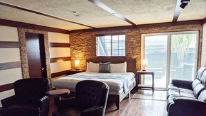 Ein Hotelzimmer mit einem Bett, zwei Sesseln, einem kleinen Tisch, einer Wand mit Steinoptik und einer Schiebetür zum Außenbereich.