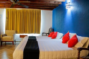 Quarto de hotel com uma cama grande, uma cadeira, um ventilador de teto de madeira e uma janela com cortinas amarelas.