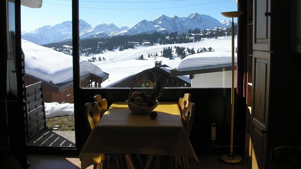 Un coin repas avec une table et des chaises, un panier de fruits et une vue sur des montagnes enneigées par la fenêtre.