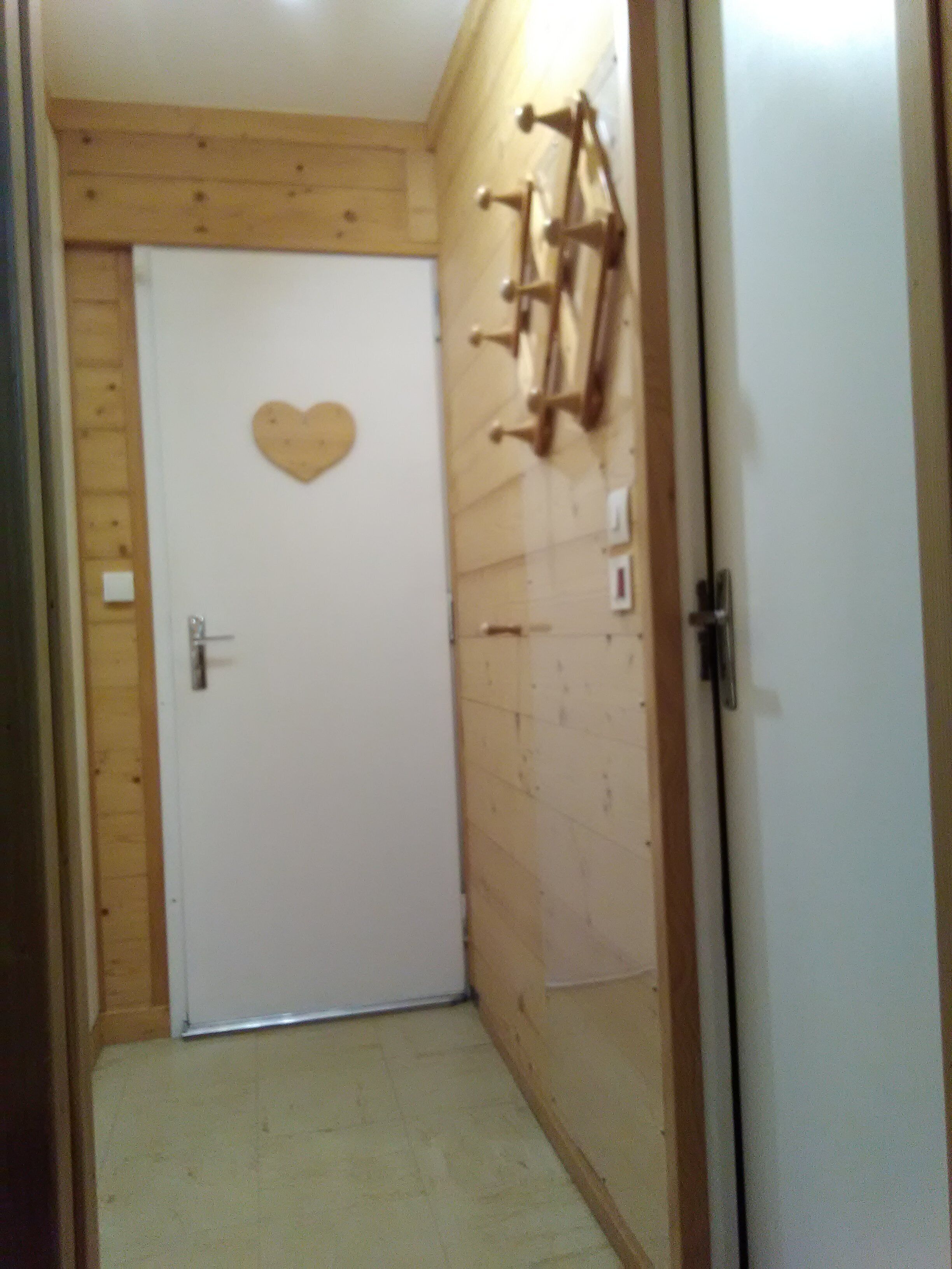 A narrow hallway with a wooden door at the end, a heart-shaped decoration on the door, and a coat rack with hooks on the wall.