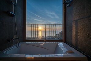 A modern bathroom with a large bathtub, a shower, and a view of the sunset over the ocean.