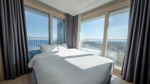 A hotel room with a large bed, two windows with curtains, and a view of the city. - Hotel the Blue Terra (Sokcho)