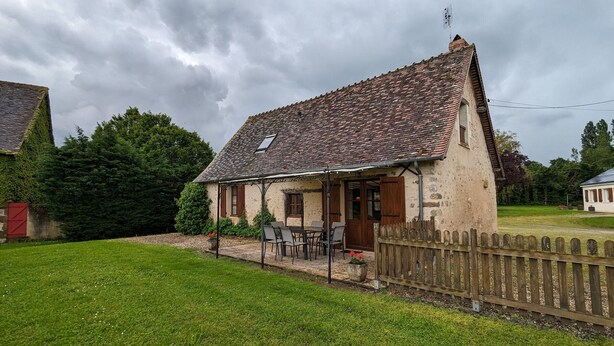 Une maison de campagne au toit recouvert de tuiles, avec un porche doté de chaises et une clôture en bois.