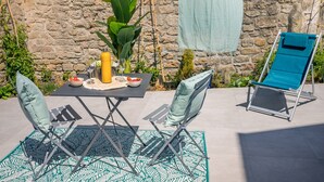 Un patio al aire libre con un muro de piedra de fondo, una mesa dispuesta con vasos y una botella, dos sillas plegables y una silla reclinable azul.