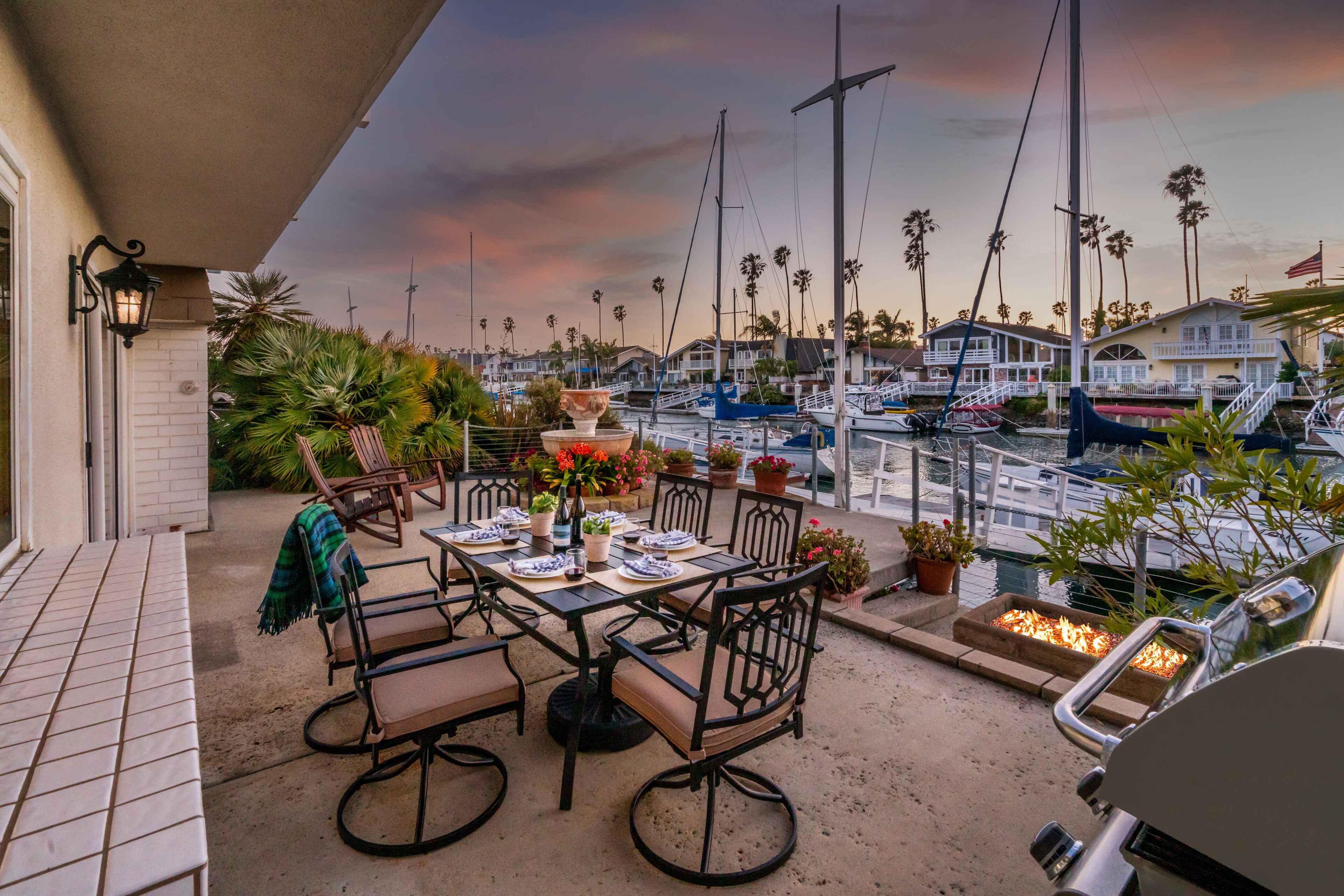 Eine Terrasse im Freien mit einem Esstisch, umgeben von Stühlen und Blick auf eine Marina mit Booten und Palmen.