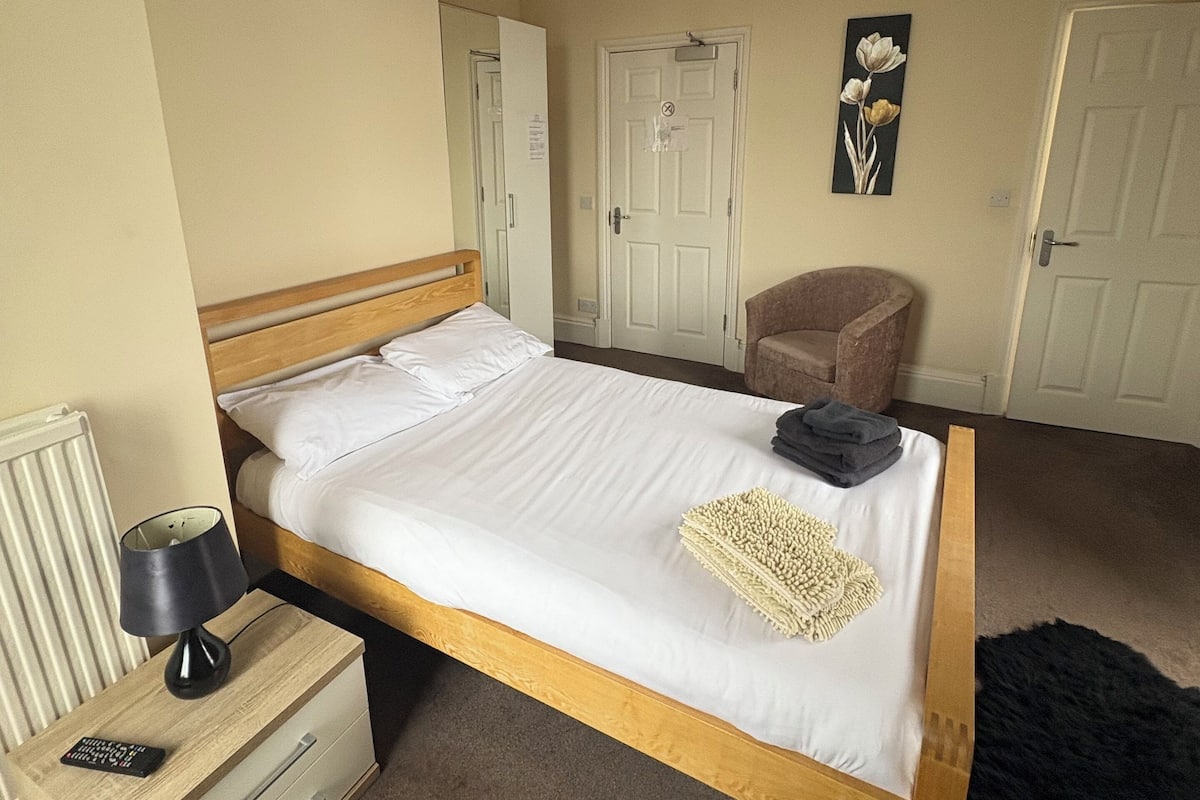 Una cama individual con estructura de madera y ropa de cama blanca, una mesita de noche con lámpara, una silla y una puerta en una habitación con paredes color beige.
