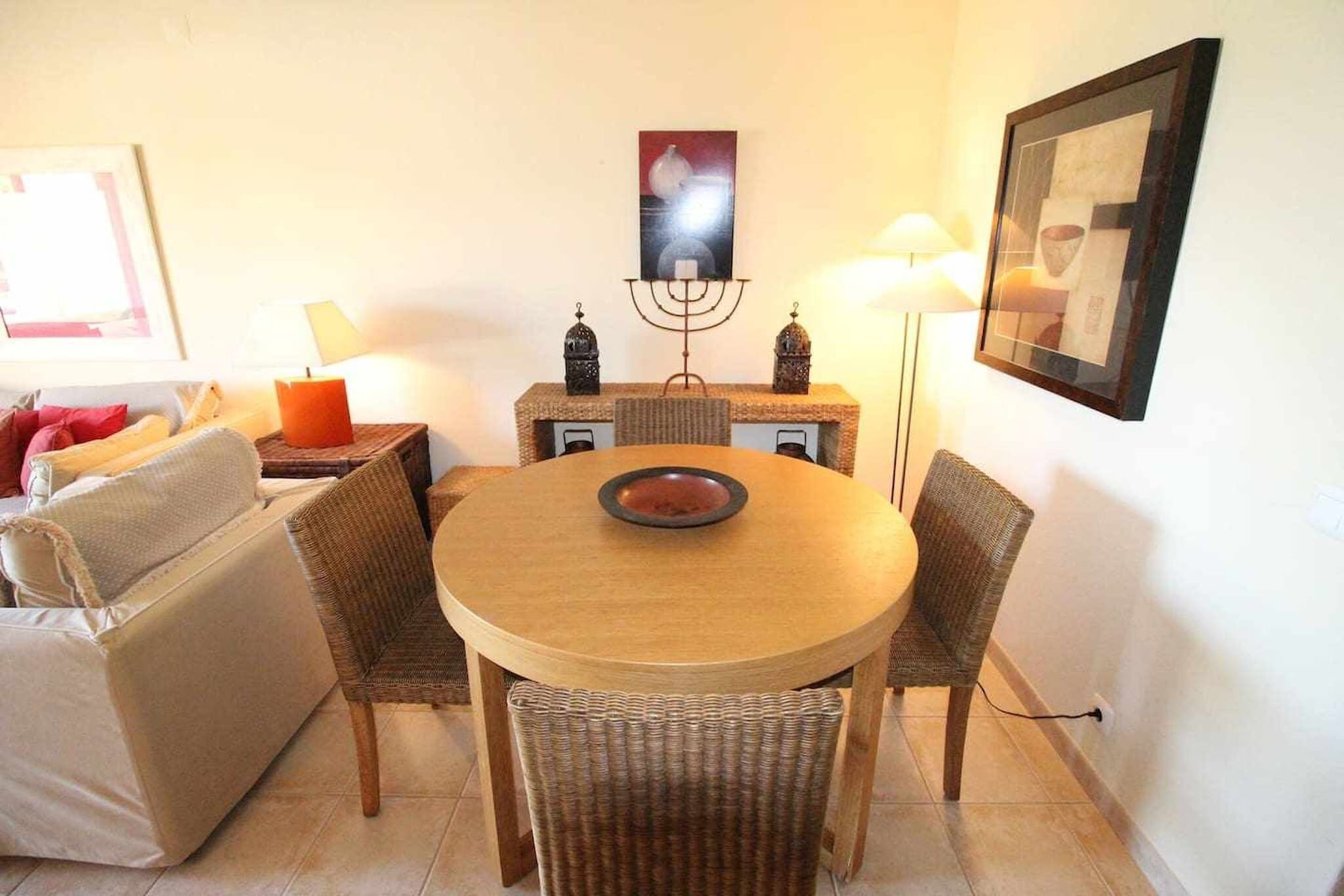 Un coin repas avec une table ronde, des chaises en osier, un chandelier et un tableau encadré au mur.