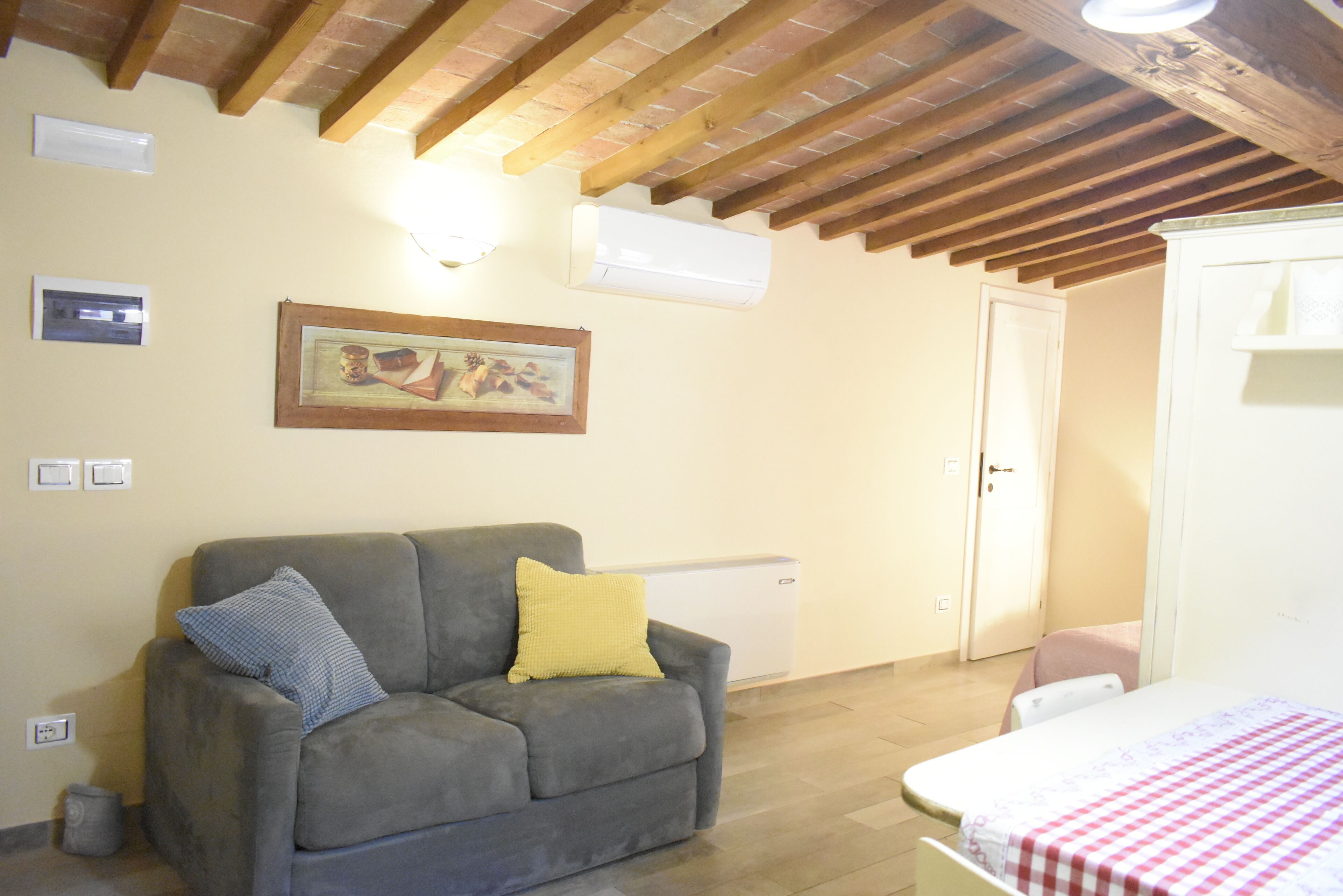 Une pièce avec un canapé gris, un lit recouvert d’une couverture à carreaux, un radiateur blanc et un plafond en bois.